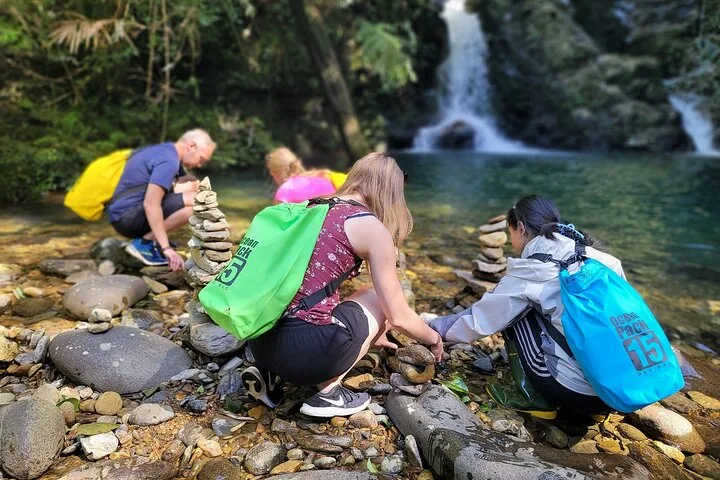 People stacking rocks near a river with a waterfall in the background.
